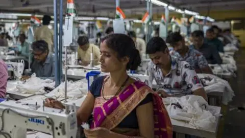 Getty Images Employees at a garment factory in India with a woman at a sewing machine in the foreground wearing a purple and gold dress
