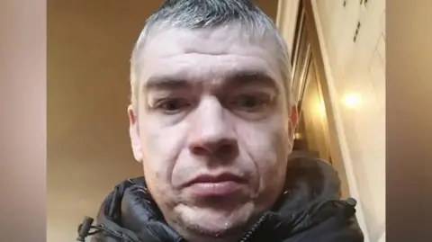 Facebook A man in his late thirties with grey hair staring from above into a camera. He is in a dimly lit room and wearing a black coat.