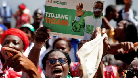 Reuters Female PDCI supporters protest against a court judgement to remove their leader Tidjane Thiam from the electoral list in Abidjan, Ivory Coast. One holds up a campaign poster of Thiam waving - 24 April 2025