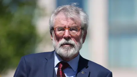 PA Media Gerry Adams, with grey hair and a grey beard, wearing a navy suit, blue shirt and red tie. He is wearing round orange glasses. The background of the image is blurred.