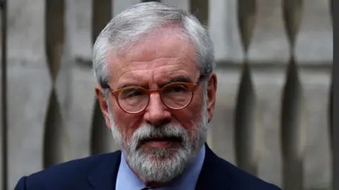 Reuters Gerry Adams has grey hair swept to the side. He is wearing red round glasses and has a beard. He is in a navy suit and blue shirt. He is looking away from the camera.