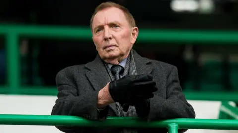 SNS Sir Tom Farmer standing in a football stand. he is wearing a greay coat and grey shirt and tie