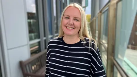OLIVIA RICHWALD/BBC A blond-haired woman wearing a blue top with narrow white stripes smiles as she stands outdoors with a blurry background of a bench and panes of glass behind her. 