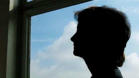 BBC A silhouette of a woman looking out of a window, with a view of blue sky and white clouds.