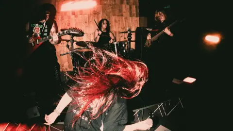 Jordan Rhys A woman with red hair is pictured rocking her head back and forth. Other band members holding guitars and playing drums can be seen in the background.