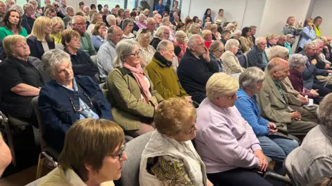 BBC A meeting room full of people sitting staring ahead. It is men and women of all ages paying attention to a speaker.