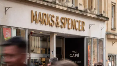Getty Images Marks & Spencer sign above a shop in gold lettering. People are walking past the shop and are blurry.