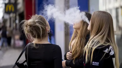 BBC Three young women with long light brown hair, the backs of their heads pictured, blow plumes of smoke in the air. They are standing in a street with a McDonalds sign in the background.