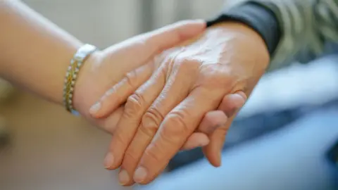 Getty Images An older person's hand is held by a younger person in a close-up shot. The younger person is wearing a gold and silver bracelet. The older person's forearm is covered by a green and black sleeve.