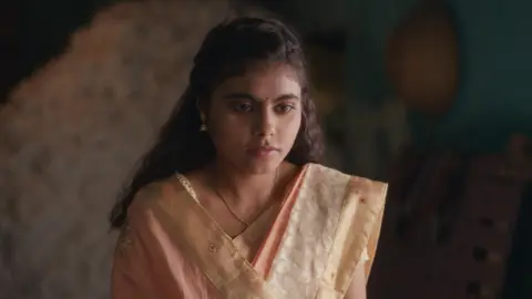 Dhun Production The image shows a young Indian girl clad in a traditional pale pink saree looking visibly upset. 