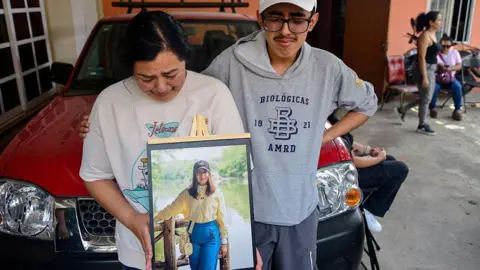 MARCO ANTONIO PEREZ/AFP via Getty Images) Relatives of América Sánchez hold up a framed photograph of the young cadet in her hometown of Xalapa. in the photo, América Sánchez, is posing in a yellow blouse and jeans in front of a lake. Her relatives are crying. 