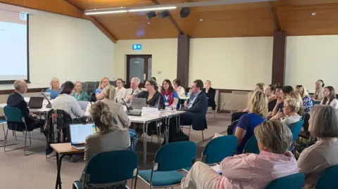 BBC Politicians sit around a table in a town-hall type setting. Members of the public look on. Tom Binet is sat centrally. 