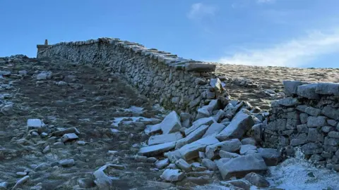 Dave Somerville A granite stone wall rises up the side of a steep mountain. A section of the wall has collapsed and a large number of stones are lying in rubble.