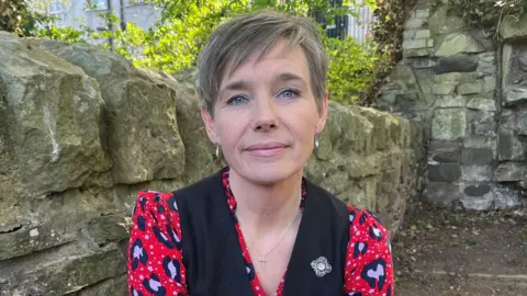 BBC Emma Bryson, with her light brown hair in a short pixie style, looks to the camera thoughtfully against a background of a stone garden wall and greenery. She wears a red, pink and black patterned shirt with a black waistcoat and a blue and gold enamel brooch.