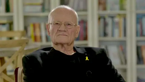 BBC Danny Yatom sits in an office, in a black shirt and jacket with glasses on, with books in the background