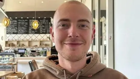 Handout Eli smiles at the camera in a cafe. He has very short haven hair, light short stubble and dark eyebrows. He wears a brown hoodie, and has a nose ring.