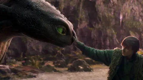 Universal Pictures A boy with swept over to the side hair, wears a green outfit, fluffy waistcoat puts his hand out and touches the nose of an animated dragon which has green eyes.
