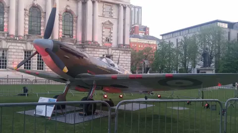 BBC A replica Supermarine Spitfire, used extensively by the Allied Forces during World War Two, on display on the grass outside Belfast City Hall, which is partially visible in the background.