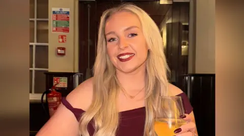 Jess Thorpe Jessica Thorpe smiling at the camera. She had long blond hair and is wearing a burgundy dress, with matching burgundy painted nails. She is holding a wine glass.