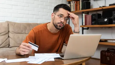 Getty Images Anxious-looking man sits behind a table in a living room, with a laptop, bills and a payment card in front of him.