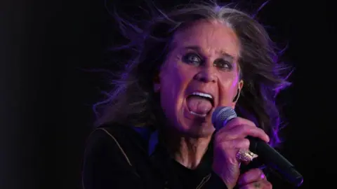 Getty Images Ozzy Osbourne is holding a microphone with his right hand and has his mouth open. The background is black. 