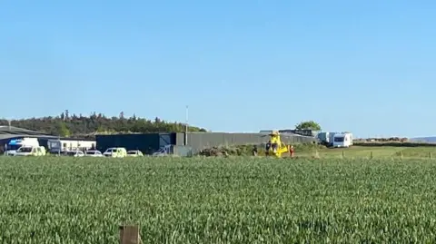 Claire Gardner Several emergency service vehicles including an ambulance helicopter in the middle of a field. Campervans can be seen in the background