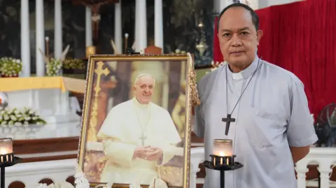 BBC/ Natalie Thomas Cardinal Pablo Virgilio David standing shirt next to a photo of Pope Francis inside a cathedral.