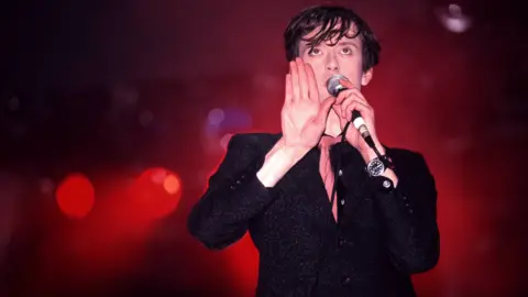 Getty Images Jarvis Cocker pictured performing in 1995. He is wearing a dark suit, black shirt and pink tie.