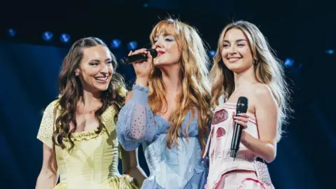 Sarah Louise Bennett Remember Monday smile and laugh on stage as they rehearse for the Eurovision song contest. The singers, in pastel shades of yellow, blue and pink, hold microphones, with Lauren (centre) the only one singing.