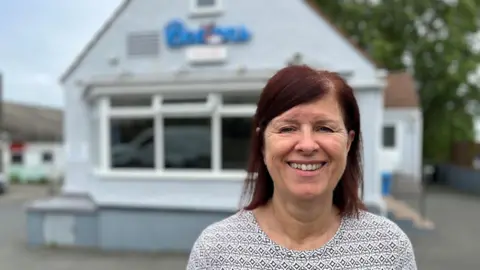 BBC Kim Ashplant smiling at looking at the camera. She is wearing a light coloured top and has dark red hair. She is stood in front of the fish and chip shop which is blurred.