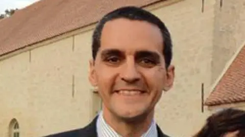 Athena Picture Agency Richard Osman, who has short dark hair and is wearing a striped shirt and suit jacket and is smiling at the camera. A beige coloured building is behind him.