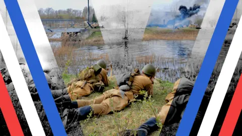 BBC A photo showing men wearing World War Two military uniforms lying on their stomachs in the grass in front of a river. On the other side of the river plumes of smoke are erupting into the cloudy sky