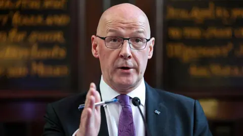 Getty Images John Swinney, who is wearing a navy blazer, a purple tie and a white shirt, addressing a press conference. Behind him, blurred, is a board with calligraphy writing on it.