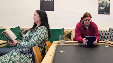 BBC Two women, sat facing away from each other, both with dark hair. One is reading a paperback book, the other a purple tablet.