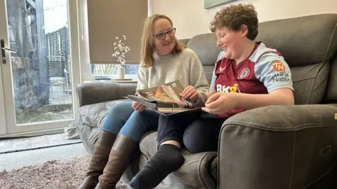 BBC Amy wears glasses, a grey jumper and long brown boots. She is sat next to her 12-year-old son Cooper, a child who has brown hair and is wearing an Aston Villa top. They are sitting on a sofa in a living room looking at a book with a French door behind them. They are both smiling.