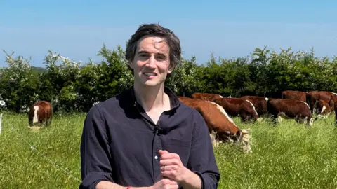 BBC Farmer Aidan Stanley stands in a field near Bath while behind him a small herd of brown and white cows is visible. The grass in the field is very tall, and there is also a hedge in the background