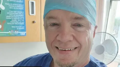 Instagram/Robert Albon Robert Albon, who appears to be in a medical facility. He has a blue top, possibly a gown, that is just visible and a blue head covering as you might see in a surgical setting. He is smiling and has a small amount of short light coloured beard growth on the angle of his chin, roughly the width of his mouth.