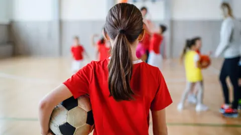 Getty Images Rear view of small girl standing with ball indoors in gym class, physical education concept.