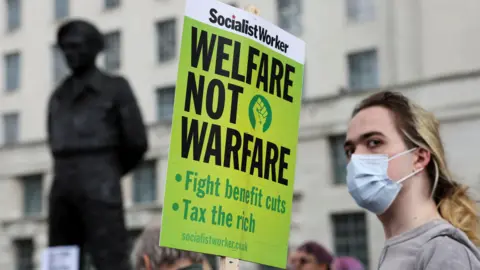 BBC A demonstrator carries a placard at a protest against possible welfare cuts, on the day British Chancellor presents the Spring Statement to Parliament, in London, Britain, March 26, 2025.
