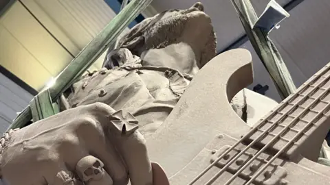 BBC A statue of a man holding a guitar, viewed from below and looking straight up to his chin. The fingers he is using to play the guitar have rings on them, including one of a skull.