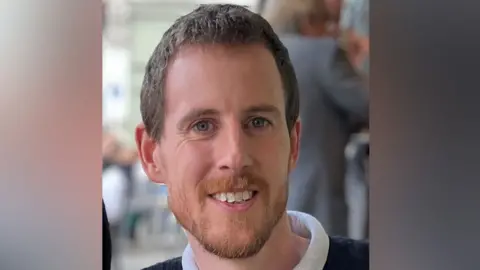 Thames Valley Police Man with short brown hair and beard smiling at the camera. He is wearing a blue sweater with a white collar