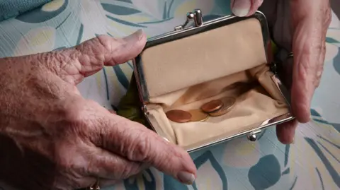 Getty Images An image of an older woman's hands holding a small purse - it is open and a handful of coins can be seen inside it. The hands and purse are resting on the woman's lap and she is wearing a teal dress or skirt with a floral pattern.
