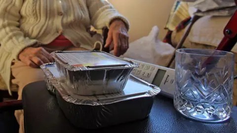 Getty Images Two foil food containers stacked, and empty glass and a landline wireless phone sit on a black leather-like surface. Behind it is an elderly person wearing a beige cable knit cardigan with buttons. Face is not visible and clothing does not make it possible to distinguish if they are male or female.