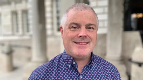 BBC Councillor Michael Long with short grey hair is smiling at the camera. He is wearing a purple shirt with a diamond pattern on it.