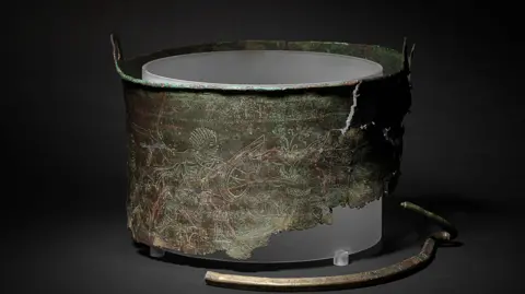 David Brunetti/National Trust A photo of the Bromeswell Bucket, which has been displayed on a black surface with a black background. Part of the bucket is missing and etchings can be seen on the bucket's surface.