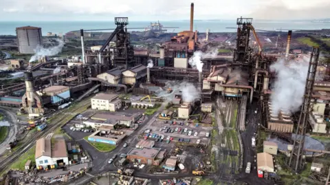 PA An aerial shot of Tata Steel's Port Talbot steelworks in south Wales. Smoke is coming out of a number of chimneys across the site, while cars are parked in the middle. To the right-hand side rail tracks lead out the works, while the sea can be seen behind it.