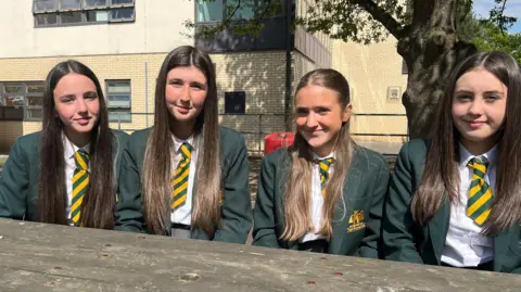 BBC Four school girls sit on a bench in front of their school. They all have long hair and are looking into camera. Their uniform is green and yellow, they wear ties and blazers.
