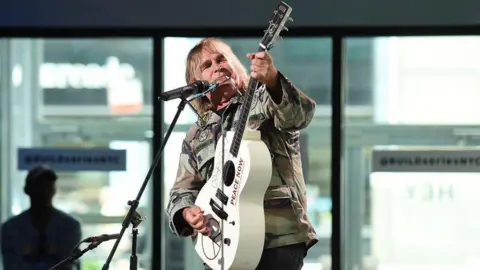 Getty Images Mike Peters who has long blonde hair and is wearing a khaki camouflage jacket. He is stood at a microphone stand and is holding a cream guitar which he is playing and looking at. Behind him are some large windows and a man wearing a baseball cap.