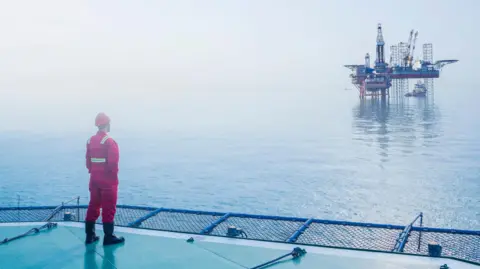 Getty Images an oil worker in a red uniform and and hard hat stands on one North Sea platform looking across the water to another platform