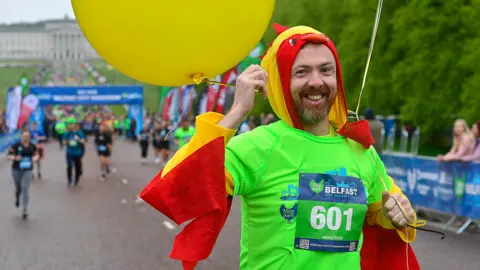 Pacemaker A man in a green t-shirt dressed in a chicken outfit at the start of the 2024 marathon. he's smiling, holding a yellow balloon and is running down Prince of Wales avenue at city hall